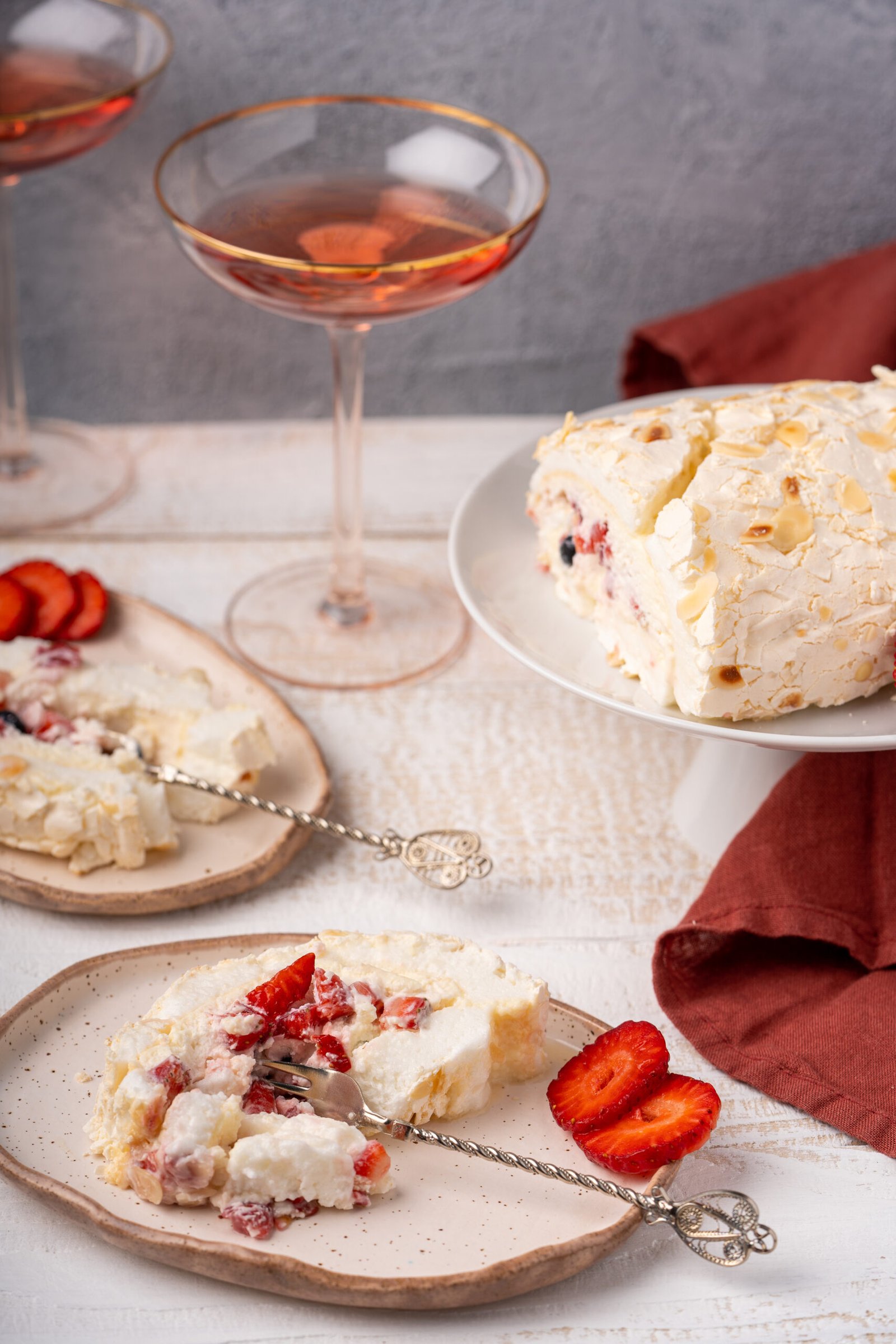 Sliced homemade meringue roulade cake with whipped cream, blueberries and strawberries on the plates and glasses of rose wine on white wood table.