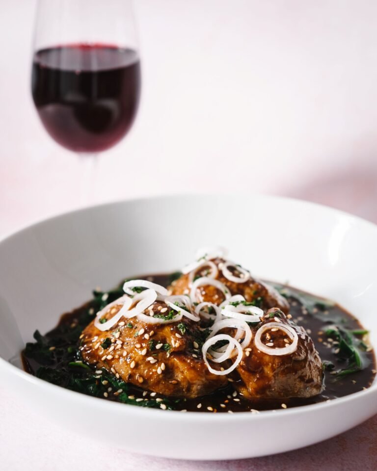 Hoisin Braised Pigs Cheeks with wilted spinach and sesame seeds served in a white bowl with a glass of red wine