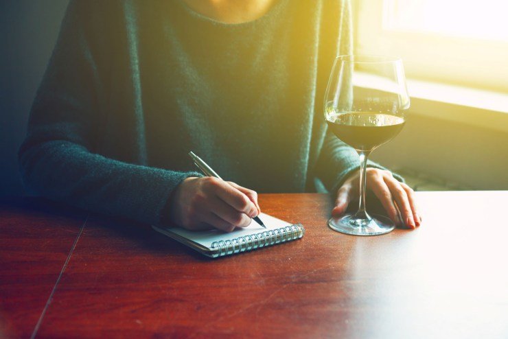 writing in a wine storage notebook with a pen on a wooden desk with a glass of red wine