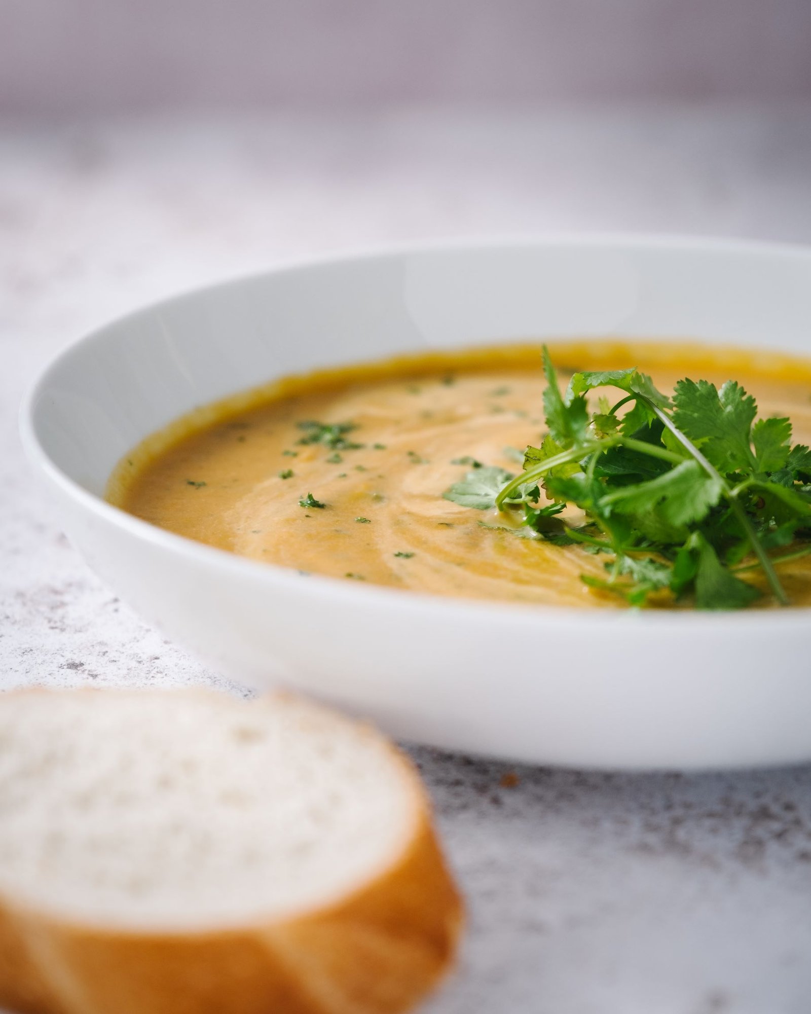 Carrot and coriander soup in a white bowl with a cilantro garnish and a slice of bread for FOOD RECIPES on Maisonbonhomie